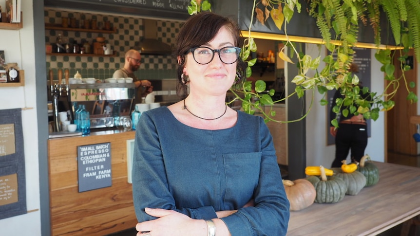 A woman in a denim dress stands in a cafe with her arms crossed, smiling.