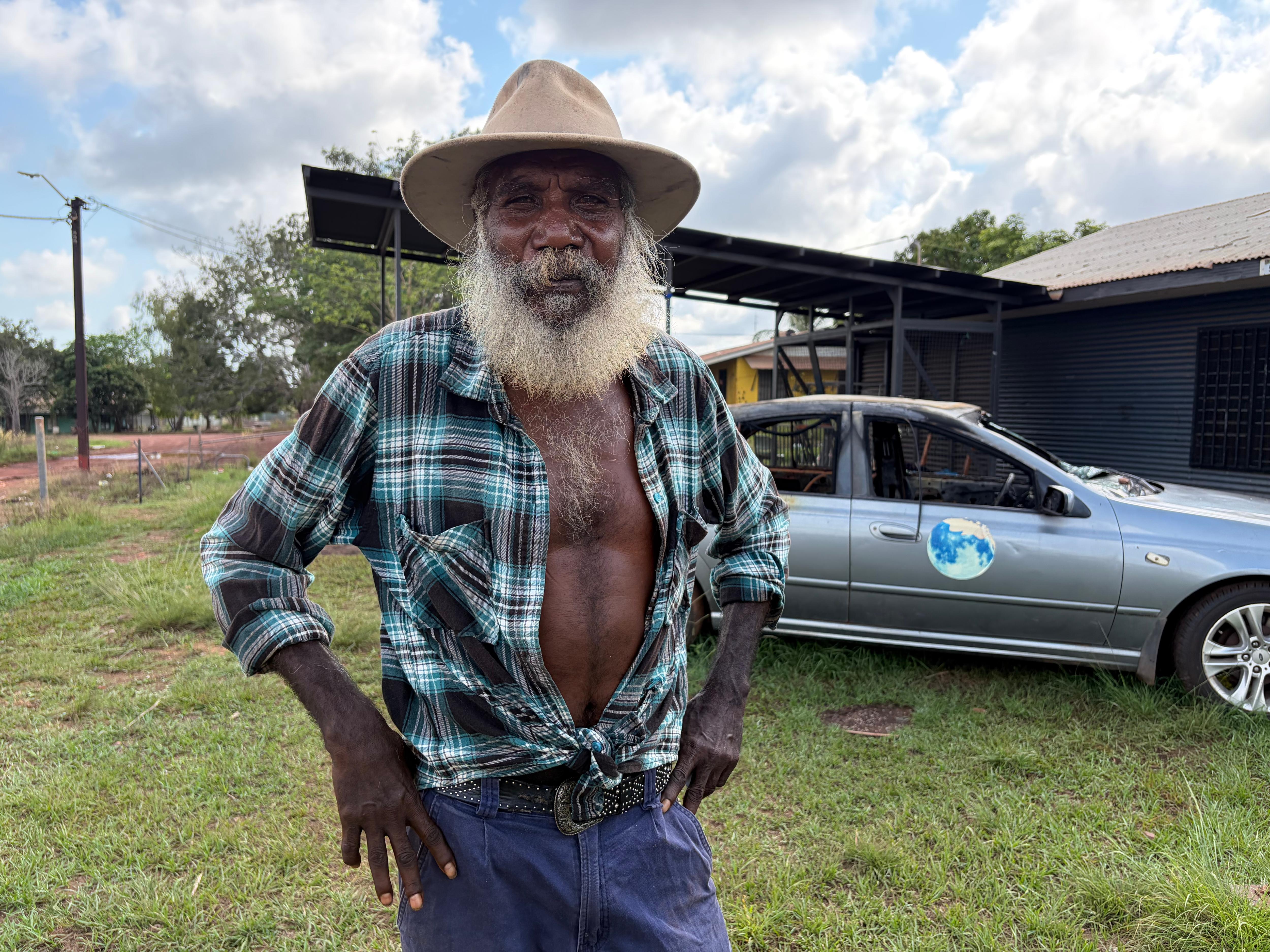 Desmond Longmair stands in front of a station wagon parked on green grass wearing a green, unbuttoned flannelette shirt.