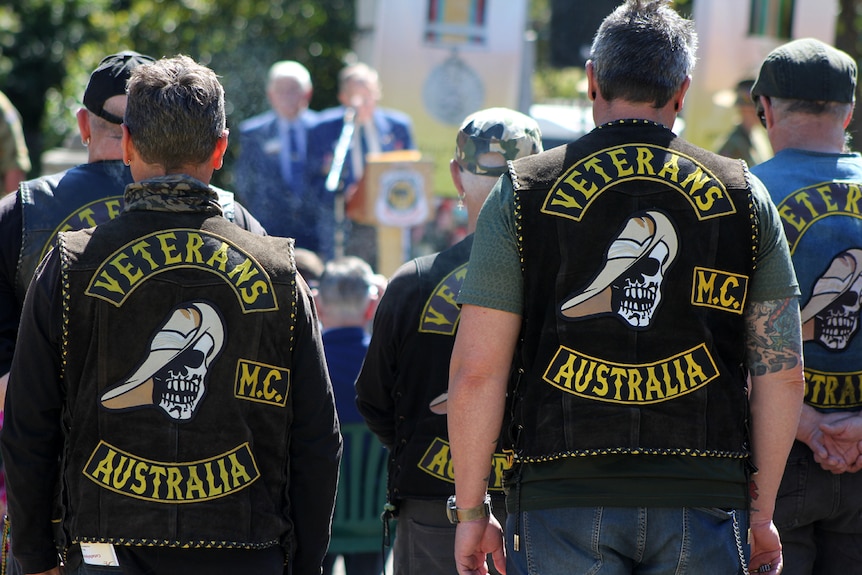 Vietnam vets stand with their jackets on, back to camera, while a memorial service takes place.