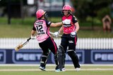 Two Sydney Sixers WBBL cricketers run to each other to celebrate their win on the pitch, with one wearing a beaming smile.