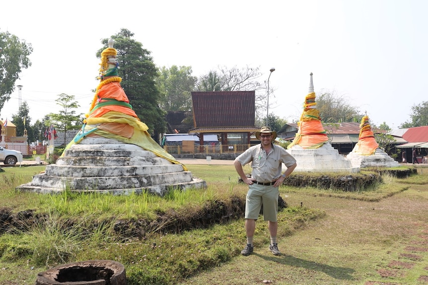 A recent photo of a man in shorts and hat standing smiling near large bell-shaped monuments in a grassy field