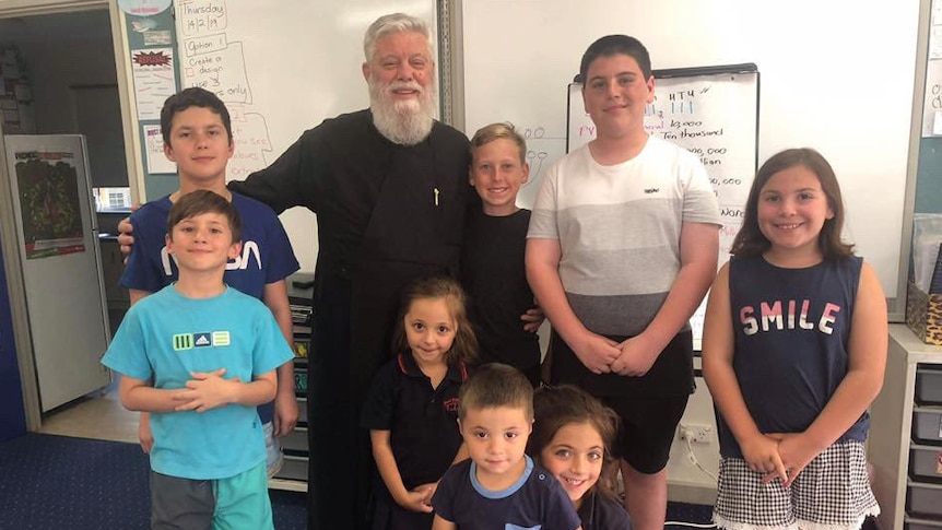 A main with a grey beard and hair wears black clothes. He is surrounded by children ranging from 5-13. They all smile. 
