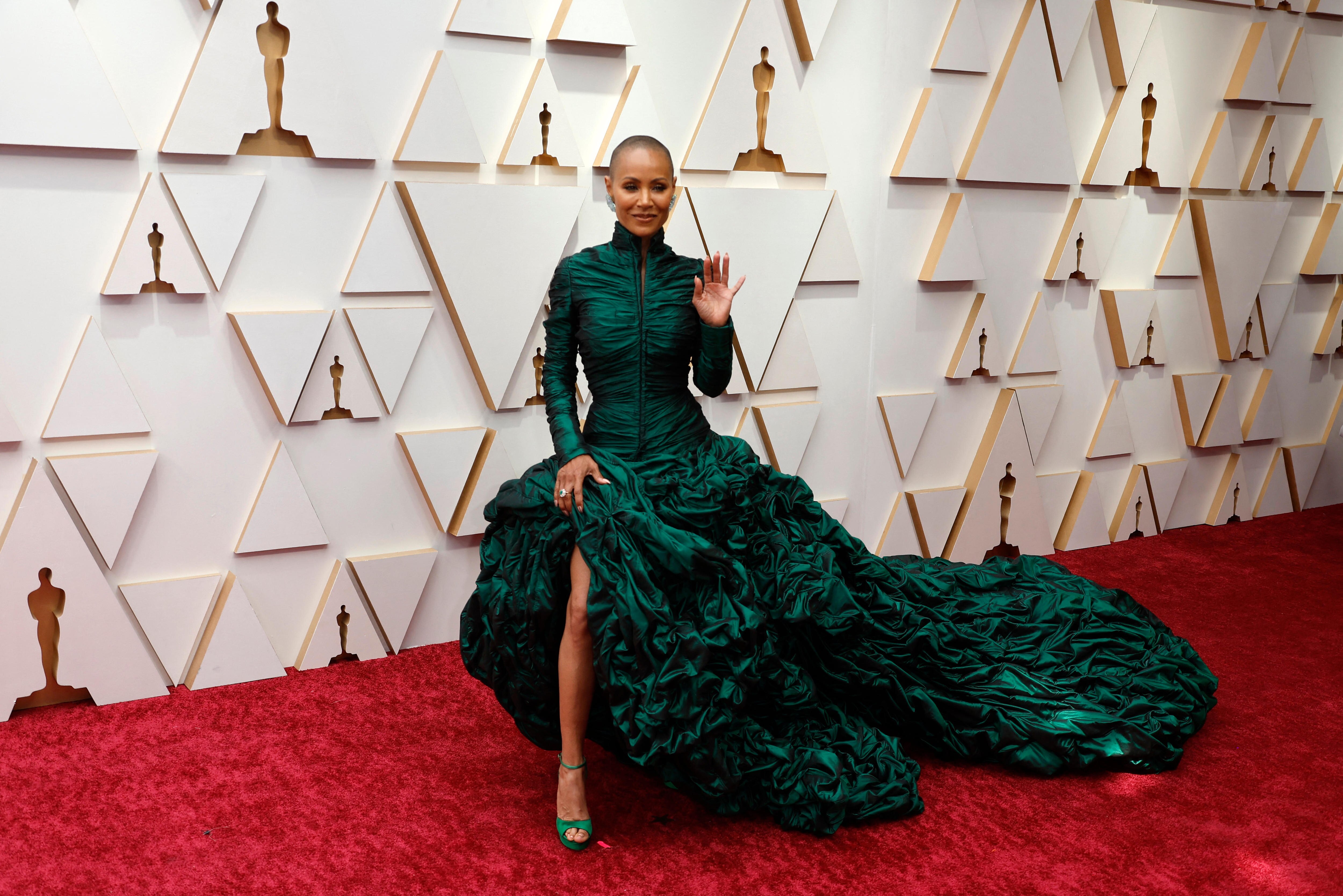 A woman wearing a dark emerald green gown with a train stands on a red carpet in front of a white media wall at the Oscars.