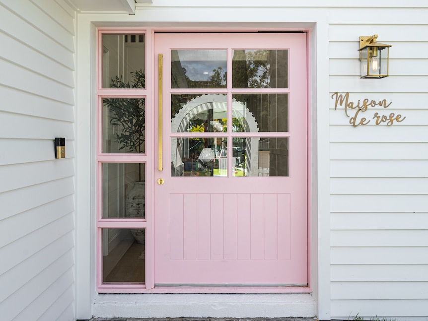 A pastel pink front door to a white multi-storey weatherboard house. 'Maison de rose' sign on the right