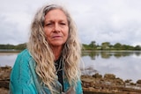 portrait of a woman, not smiling, long grey curly hair in a blue shawl by a glassy river  