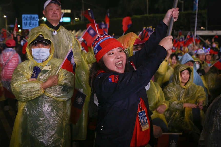 A woman wearing a red beanie smiles as she holds up a flagpole, in a crowd of people in plastic ponchos