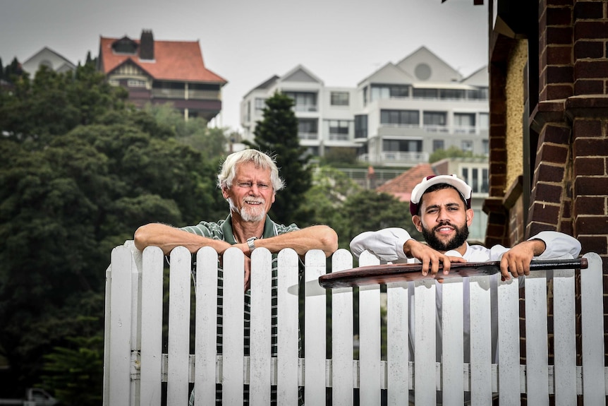 Two men peaking over a white picket fence