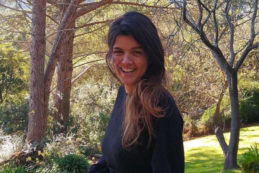 A smiling woman stands on a deck in front of a lush, green garden