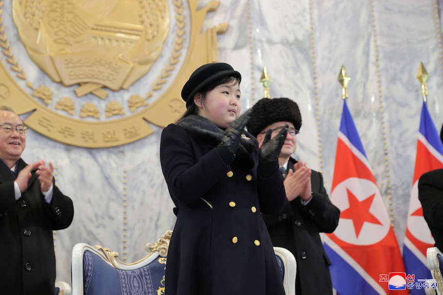 A young Korean girl in dark winter clothing stands and applauds in front of a large golden seal and North Korean flags.