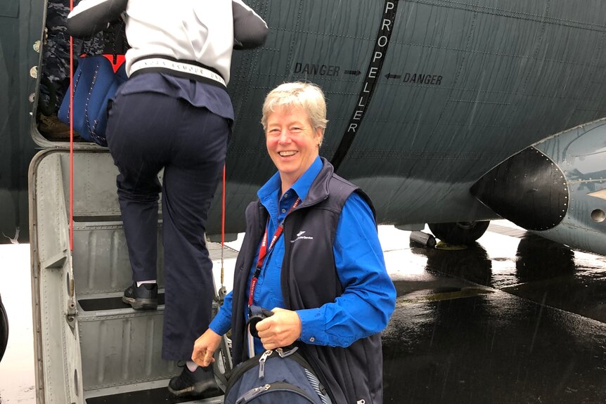 Cath walker stands in front of plane stairs as a woman in the background is entering the plane