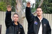 Two men in black jackets give a white power salute in a suburban backyard.