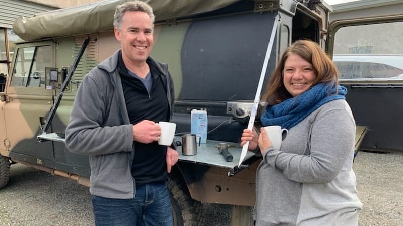 A man and woman stand in front of a Land Rover drinking coffee.