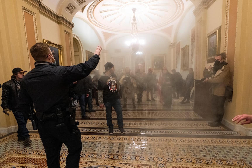 an officer in the foreground holds out his hand against advancing rioters, as smoke fills the lobby