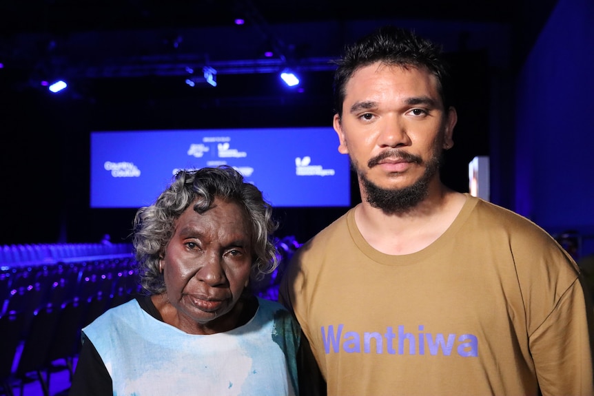 An older Indigenous woman standing next to a young Indigenous man with a big screen and blue spotlights in the background 