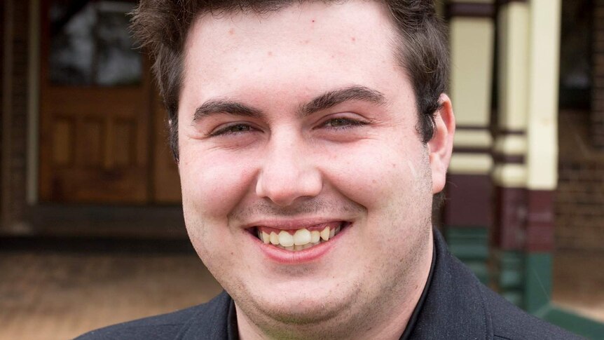 Close up photograph of a young man standing in front of a university building.