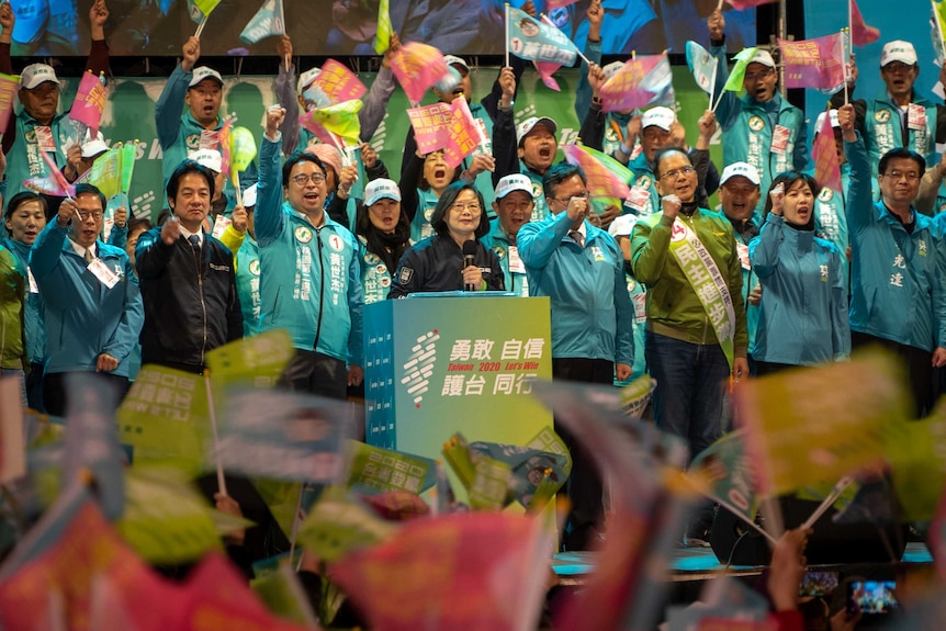 Tsai Ing-wen holds a microphone and speaks while supporters hold flags and cheer.