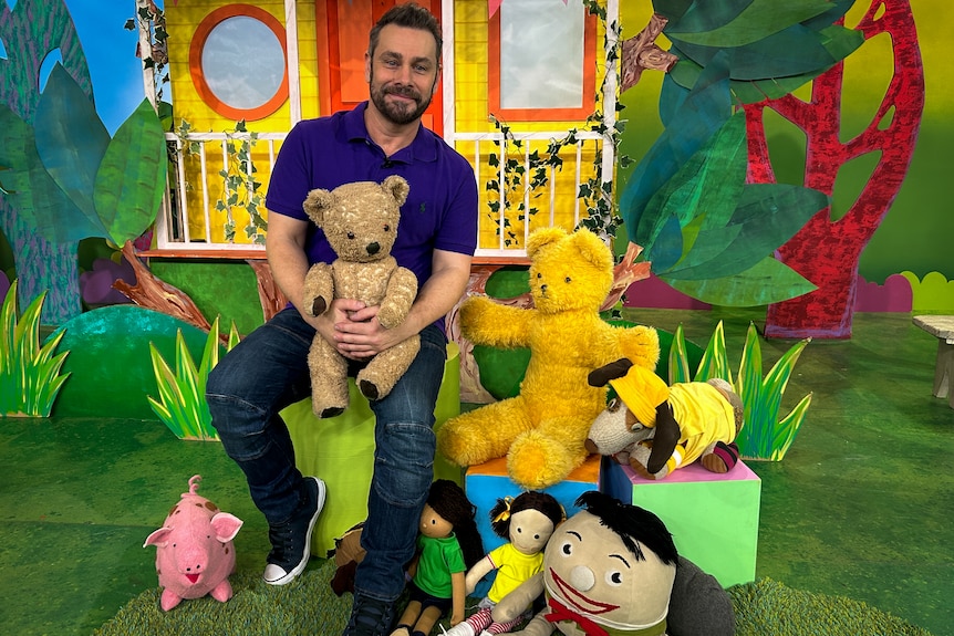 A man sits holding a teddy bear on his lap surrounded by toys in front of a colourful paper house. 