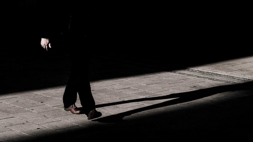 Black and white photo (except for brown shoes) of man in suit, top half obscured by darkness, and his shadow