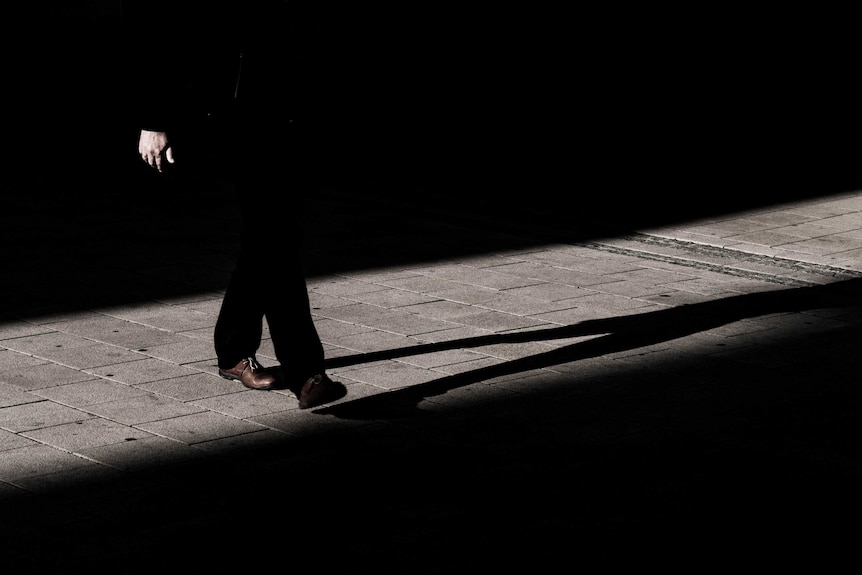 Black and white photo (except for brown shoes) of man in suit, top half obscured by darkness, and his shadow