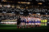 Players, umpires and fans, stand during an Anzac Observance before a night AFL match