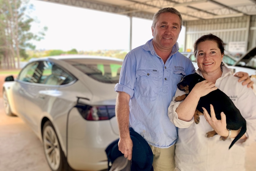 Man and woman holding black puppy standing in shed in front of their electric car