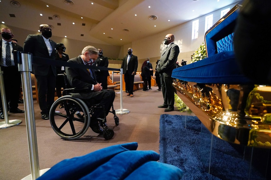 A man in a wheelchair bows his head in front of a gold casket with blue lining. Several people stand behind him.