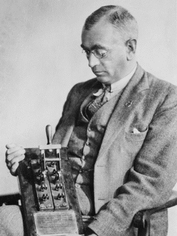 A man wearing glasses and a suit, tie and waistcoat, sits in a chair holding a large electrical switch.