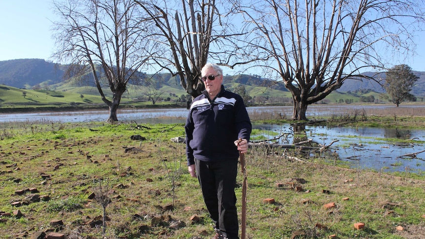 A 70 year old man stands in paddock surrounded by hills and water holding a rusted bed leg.