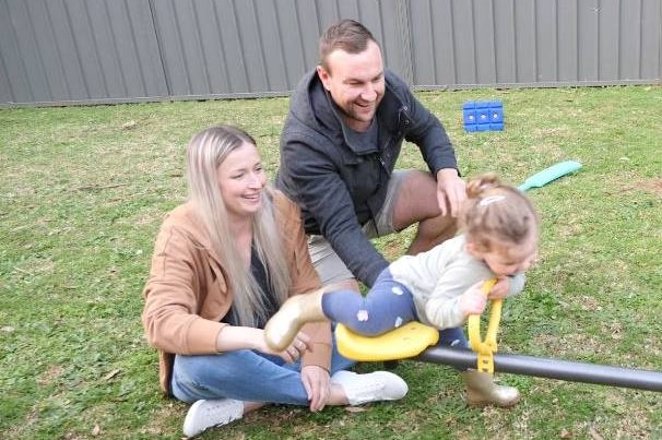 Man and his wife sit on the grass with their little girl on the seesaw smiling.