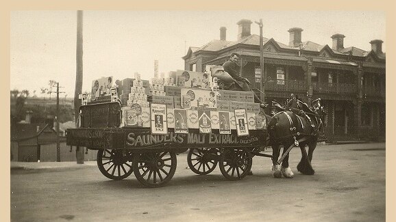 A man atop a horse and cart, loaded with boxes and cans and signs for Saunders Malt Extract.