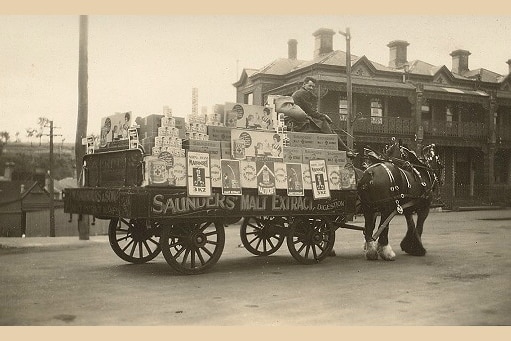 A man atop a horse and cart, loaded with boxes and cans and signs for Saunders Malt Extract.