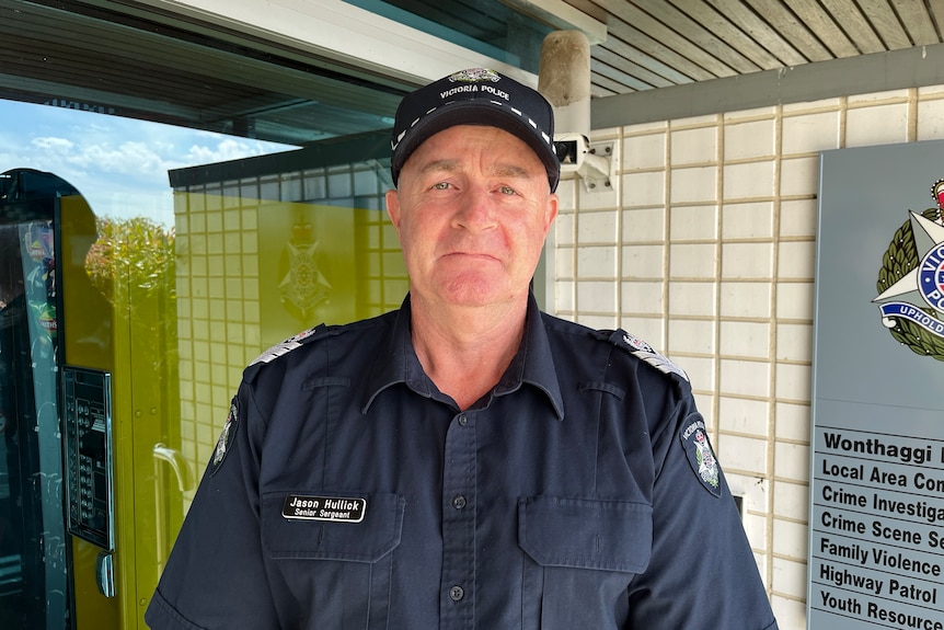 A police officer, wearing a cap, stands in front of a brick police station.