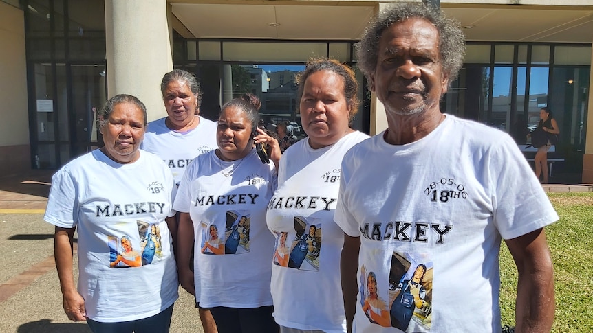 A group of Indigenous men and women in matching memorial white tee-shirt with Mackey , photos and a date printed.