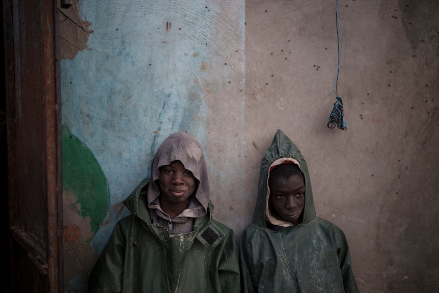 Two young boys are pictured wearing dark green raincoat while leaning against a wall.