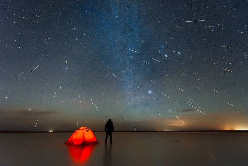 Silhouette of a person standing on lakebed looking up at sky filled with meteors.