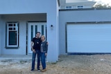 a husband and wife stand arm-in-arm with bleak expressions in front of an unfinished home 