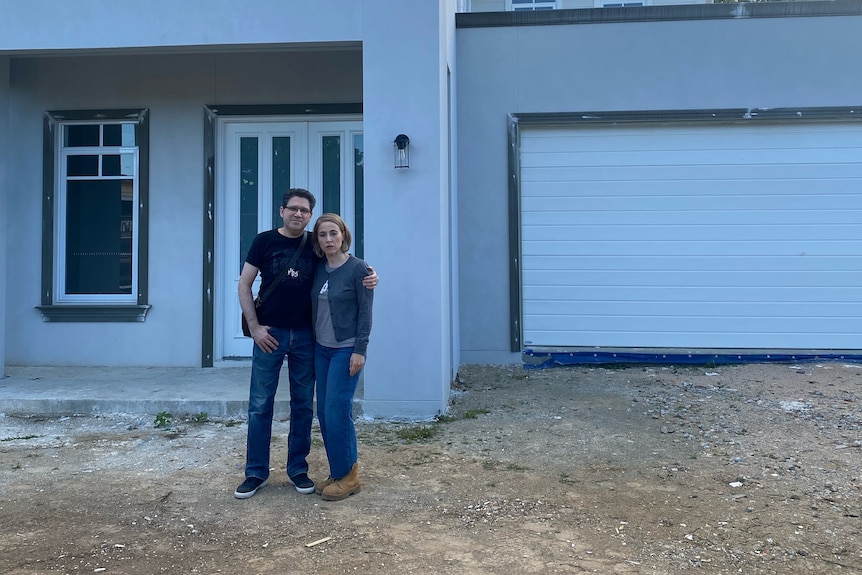 a husband and wife stand arm-in-arm with bleak expressions in front of an unfinished home 