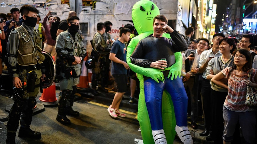 A man dressed as a cartoon-like alien holds a beer can standing in the middle of a crowd flanked by Hong Kong policemen.