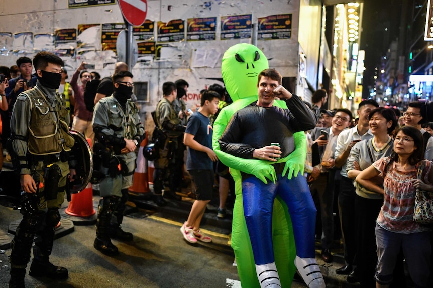A man dressed as a cartoon-like alien holds a beer can standing in the middle of a crowd flanked by Hong Kong policemen.