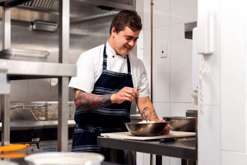 A chef works in a restaurant kitchen wearing a blue apron and white chef's shirt and leaning over a silver bowl.