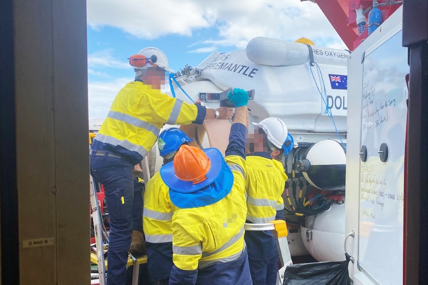 A group of men repair a large device on a boat