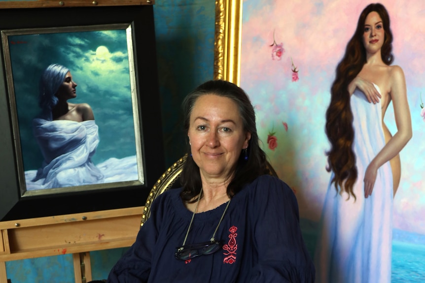 Brunette woman wearing navy shirt sitting in front of two paintings of silk-draped woman
