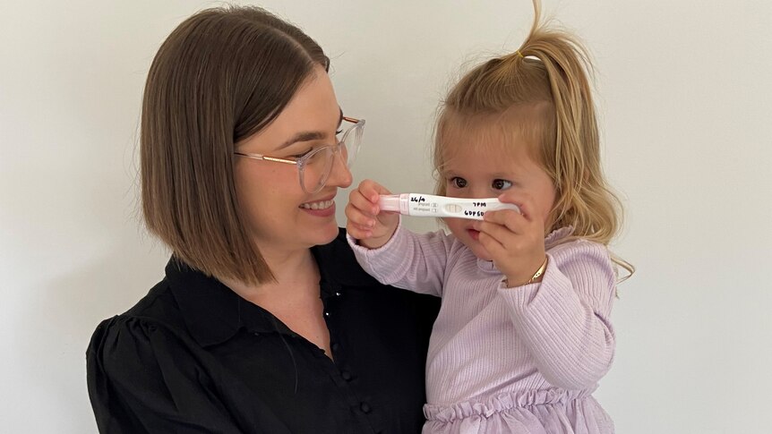 A young woman holds a two-year-old girl in her arms, with her child holding a positive pregnancy test in her hands.