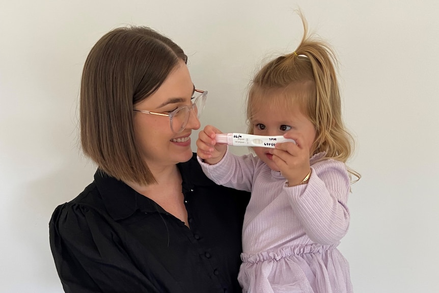 A young woman holds a two-year-old girl in her arms, with her child holding a positive pregnancy test in her hands.