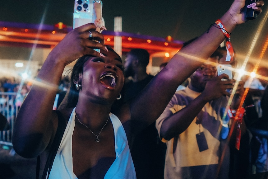 Una mujer negra con los brazos en el aire sosteniendo un teléfono móvil, con la boca abierta en celebración, top blanco