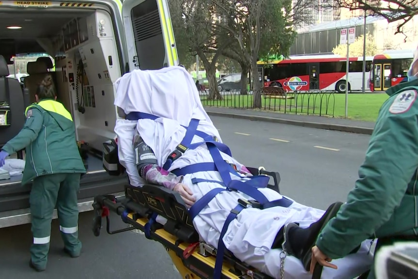 a person on a stretcher with their face covered behind an ambulance with the doors open and a paramedic looking inside