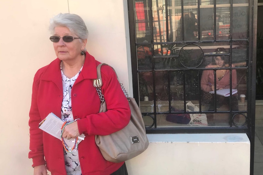 A pro-life woman stands outside an abortion clinic holding rosary beads, a clinic defender sits inside