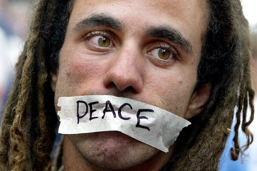 Man with dreadlocks has masking tape across his mouth with "No War" written in black marker