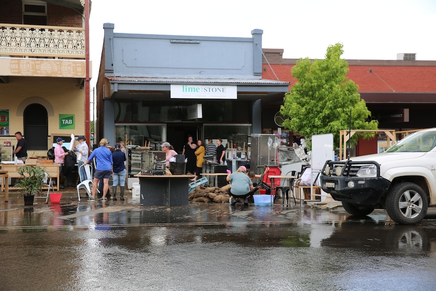 People stand on the water-soaked road in front of stores damaged by flood water.
