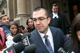Faruk Orman, wearing glasses and a suit, walks out of court as he is filmed and photographed by media.